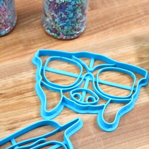 May include: Two blue plastic cookie cutters shaped like a dog wearing sunglasses. The cutters are on a wooden surface.