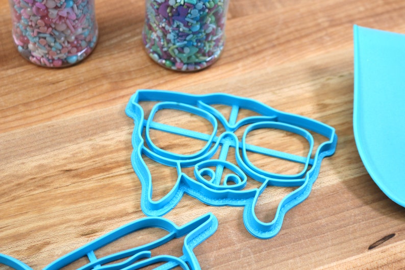 May include: Two blue plastic cookie cutters shaped like a dog wearing sunglasses. The cutters are on a wooden surface.