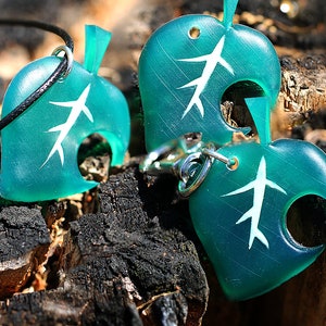 May include: Two teal green leaf-shaped charms with a white tree design. One charm is on a black cord necklace and the other is on a silver key chain.