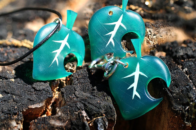 May include: Two teal green leaf-shaped charms with a white tree design. One charm is on a black cord necklace and the other is on a silver key chain.