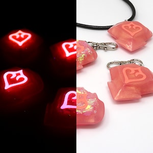 May include: Four pink resin keycaps with a white heart design. The keycaps glow pink in the dark. One keycap is attached to a black cord and two silver keychains.