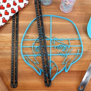 May include: A blue plastic cookie cutter shaped like a cartoon face with a grid pattern overlayed on top. The cookie cutter is on a wooden surface with a rolling pin, a red and white oven mitt, and a blue spatula.