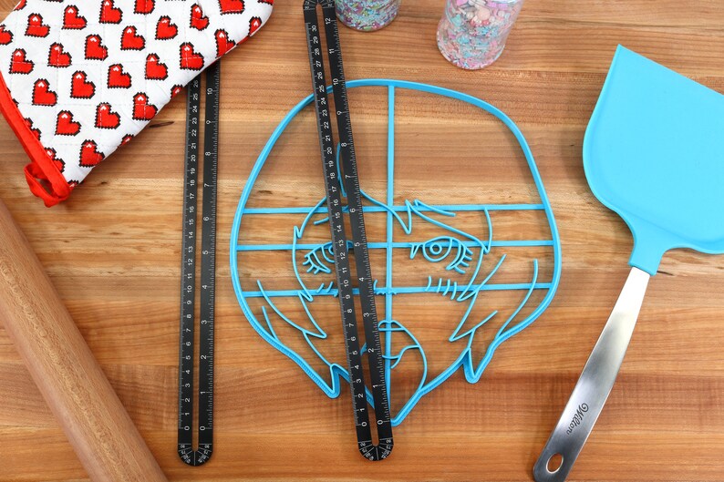 May include: A blue plastic cookie cutter shaped like a cartoon face with a grid pattern overlayed on top. The cookie cutter is on a wooden surface with a rolling pin, a red and white oven mitt, and a blue spatula.