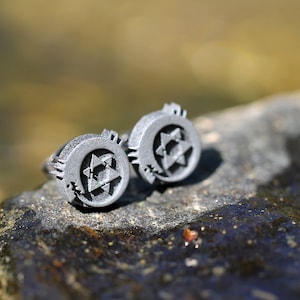 May include: A pair of silver cufflinks with a star of David design. The cufflinks are on a gray rock.