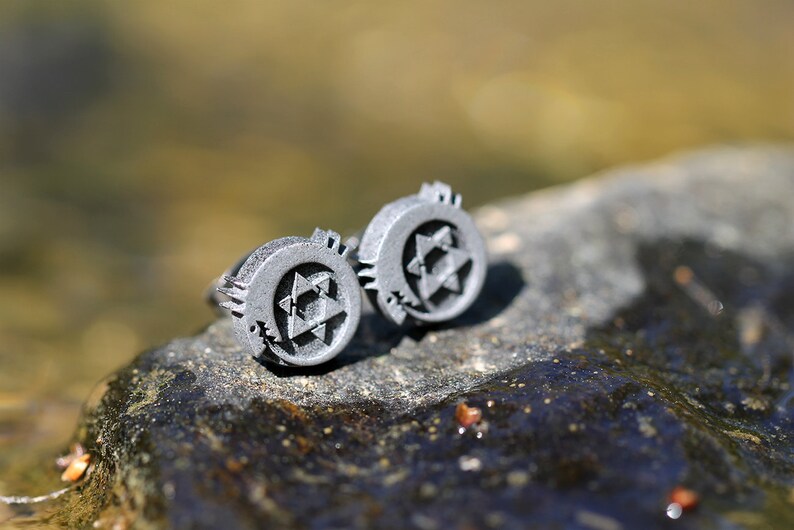 May include: A pair of silver cufflinks with a star of David design. The cufflinks are on a gray rock.