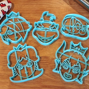 May include: Five blue plastic cookie cutters shaped like cartoon characters. The characters are wearing crowns and have different facial expressions.