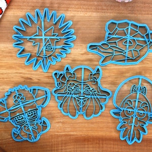 May include: Five blue plastic cookie cutters shaped like cartoon characters. The cutters are on a wooden surface.