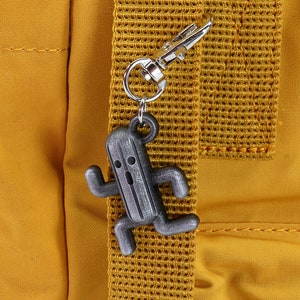 May include: A silver-colored keychain featuring a running character from the Final Fantasy video game series. The character is a gray, humanoid cactus with a surprised expression. The keychain is attached to a yellow backpack.