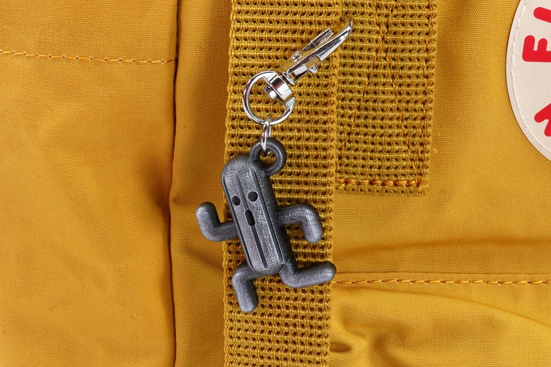 May include: A silver-colored keychain featuring a running character from the Final Fantasy video game series. The character is a gray, humanoid cactus with a surprised expression. The keychain is attached to a yellow backpack.
