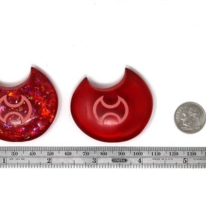 May include: Two red resin crescent moon charms with a pink symbol in the center. The charm on the left has a glittery finish. The charm on the right has a smooth finish. A ruler and a US quarter are included for scale.