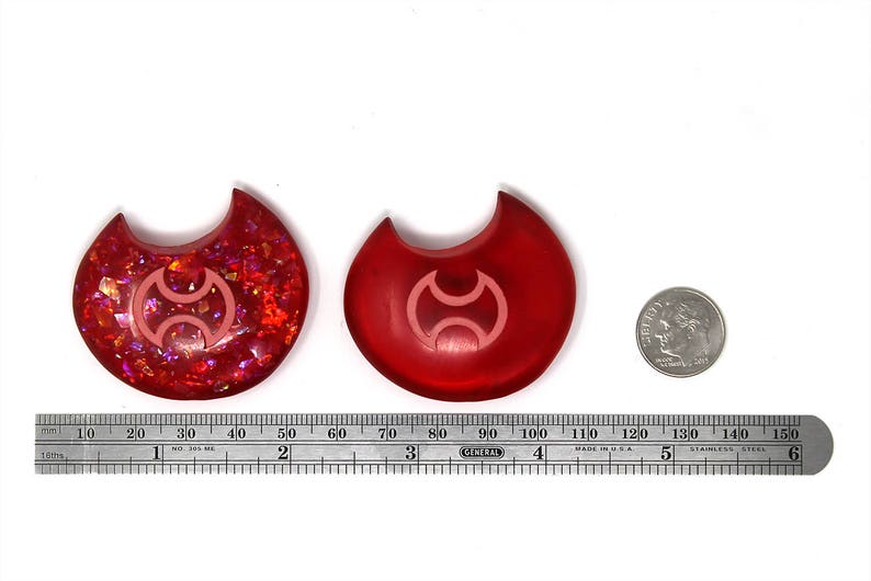 May include: Two red resin crescent moon charms with a pink symbol in the center. The charm on the left has a glittery finish. The charm on the right has a smooth finish. A ruler and a US quarter are included for scale.