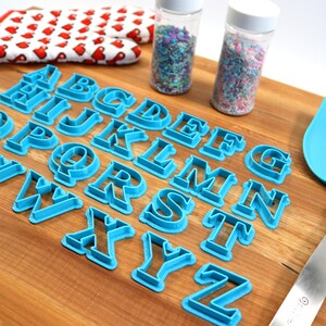 May include: A set of blue plastic alphabet cookie cutters on a wooden cutting board. There are two jars of sprinkles in the background.