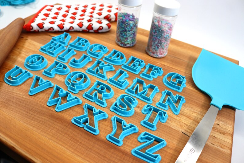 May include: A set of blue plastic alphabet cookie cutters on a wooden cutting board. There are two jars of sprinkles in the background.