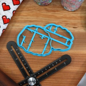 May include: A blue plastic cookie cutter shaped like a pair of lungs. The cutter is on a wooden surface with a ruler, rolling pin, and spatula.