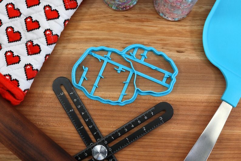 May include: A blue plastic cookie cutter shaped like a pair of lungs. The cutter is on a wooden surface with a ruler, rolling pin, and spatula.