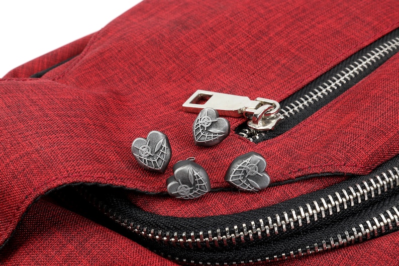 May include: Close-up of a red fabric bag with a black zipper and four silver heart-shaped zipper pulls. Each heart has a web design.