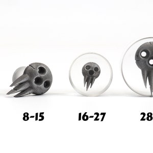May include: Four gray skull-shaped plugs in varying sizes, with the sizes labeled 3-7, 8-15, 16-27, and 28-35. The plugs have three circular eye holes and a jagged, pointed lower section. Two plugs are set in clear acrylic.
