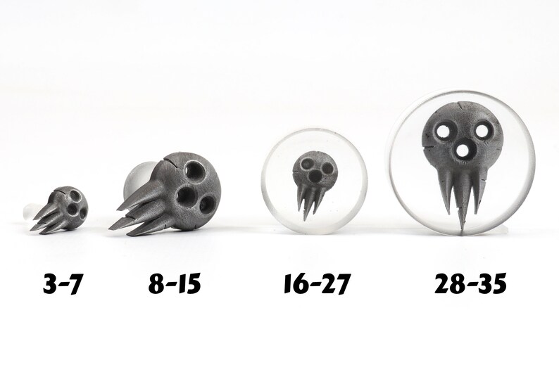 May include: Four gray skull-shaped plugs in varying sizes, with the sizes labeled 3-7, 8-15, 16-27, and 28-35. The plugs have three circular eye holes and a jagged, pointed lower section. Two plugs are set in clear acrylic.