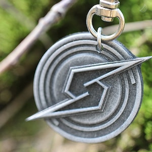 May include: A silver metal keychain with a stylized lightning bolt design in a circle. The keychain has a silver metal ring.