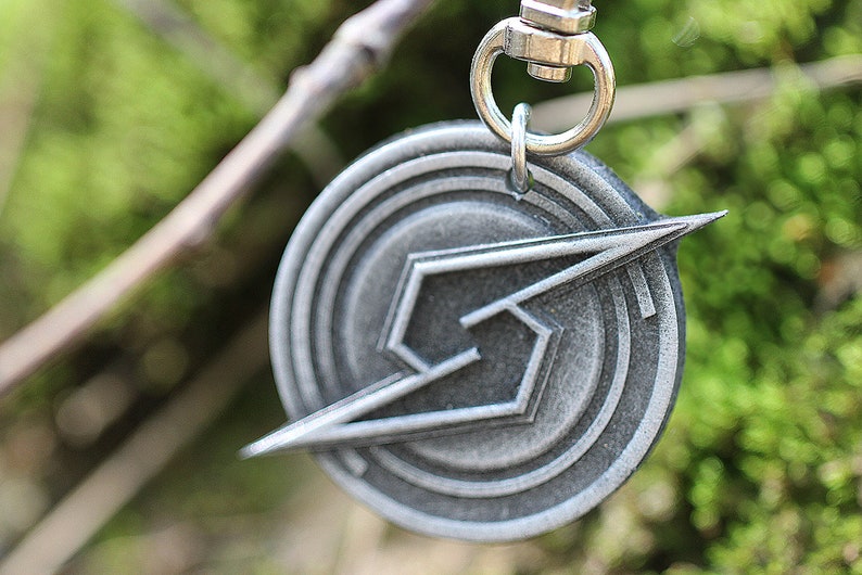 May include: A silver metal keychain with a stylized lightning bolt design in a circle. The keychain has a silver metal ring.