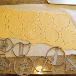 May include: A sheet of cookie dough with various shapes cut out using cookie cutters. The cookie cutters are shown on a counter next to the dough.