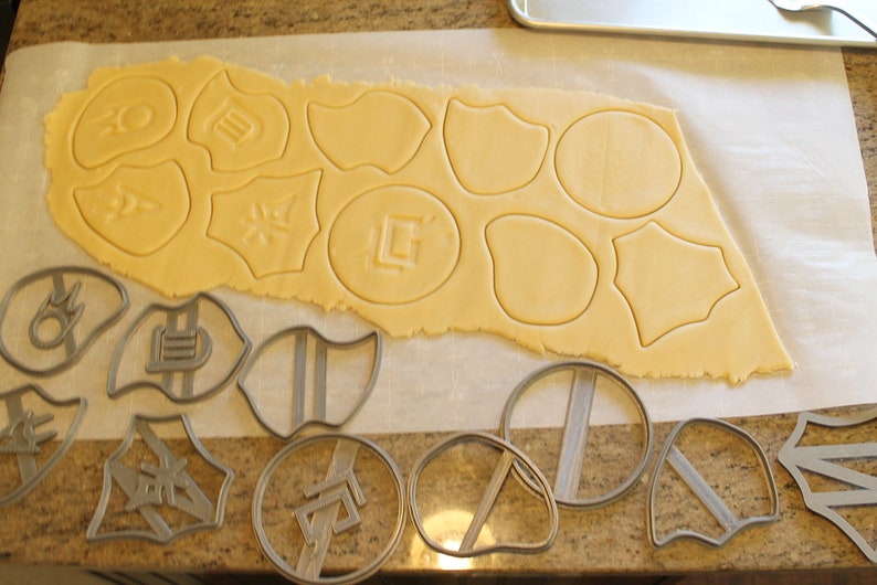May include: A sheet of cookie dough with various shapes cut out using cookie cutters. The cookie cutters are shown on a counter next to the dough.