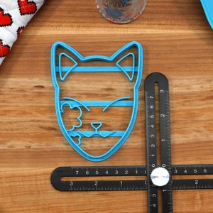 K&ouml;nnte beinhalten: Ein blauer Plastik-Ausstecher in Form eines Katzenkopfes mit geschlossenen Augen und einem Blumendesign auf einer Seite. Der Ausstecher liegt auf einer Holzoberfl&auml;che mit einem Lineal.
