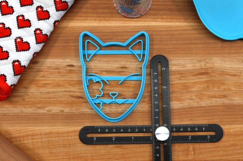 K&ouml;nnte beinhalten: Ein blauer Plastik-Ausstecher in Form eines Katzenkopfes mit geschlossenen Augen und einem Blumendesign auf einer Seite. Der Ausstecher liegt auf einer Holzoberfl&auml;che mit einem Lineal.