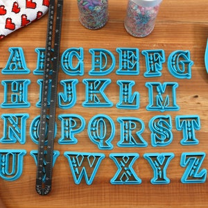 May include: A set of 26 blue plastic alphabet cookie cutters with black outlines. The cutters are arranged in alphabetical order on a wooden surface.