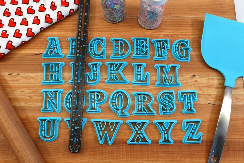 May include: A set of 26 blue plastic alphabet cookie cutters with black outlines. The cutters are arranged in alphabetical order on a wooden surface.