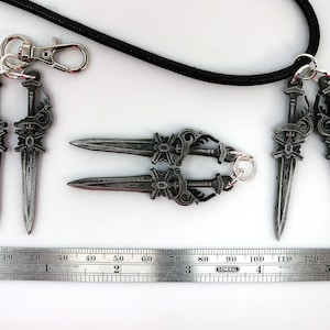 May include: A collection of silver-colored dagger-shaped pendants and keychains. The pendants are connected to a black cord and a metal clasp. A ruler shows the pendants are approximately 5 inches long.