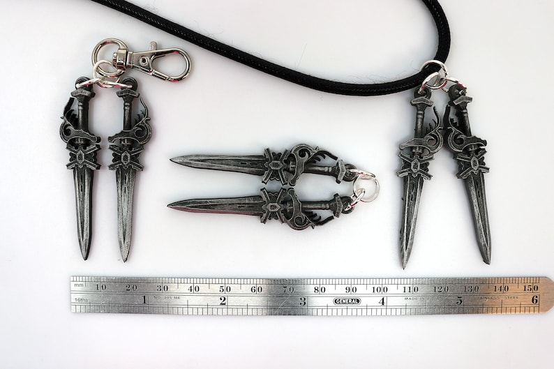 May include: A collection of silver-colored dagger-shaped pendants and keychains. The pendants are connected to a black cord and a metal clasp. A ruler shows the pendants are approximately 5 inches long.