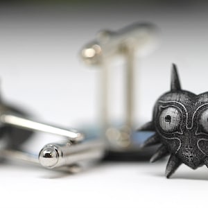 May include: A pair of cufflinks featuring a silver-coloured, detailed design of the Majora's Mask from the Legend of Zelda video game series. The mask is a heart shape with a menacing expression and sharp, pointed edges.