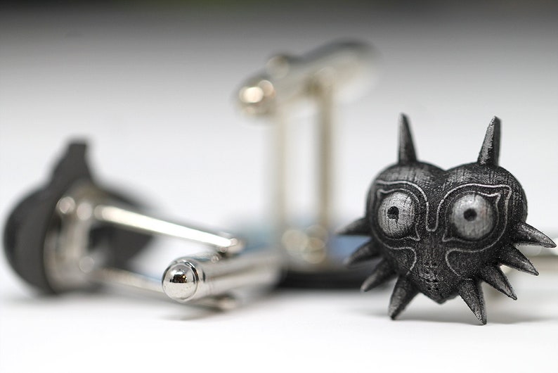 May include: A pair of cufflinks featuring a silver-coloured, detailed design of the Majora's Mask from the Legend of Zelda video game series. The mask is a heart shape with a menacing expression and sharp, pointed edges.