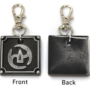 May include: A silver metal keychain with a black square charm featuring a stylised silver design. The back of the charm is plain black. The keychain has a silver clip.