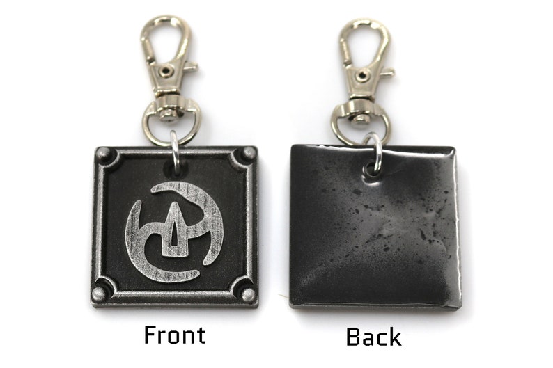 May include: A silver metal keychain with a black square charm featuring a stylised silver design. The back of the charm is plain black. The keychain has a silver clip.