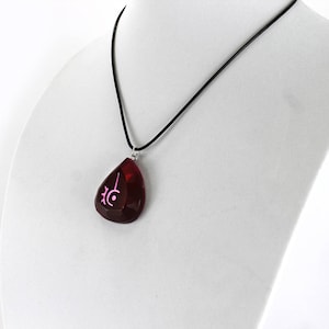 Op de afbeelding: Een ketting met een rode, traanvormige hanger met een roze symbool erop. De ketting is aan een zwart koord.