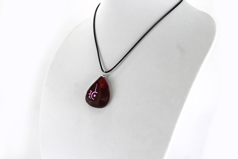 Op de afbeelding: Een ketting met een rode, traanvormige hanger met een roze symbool erop. De ketting is aan een zwart koord.