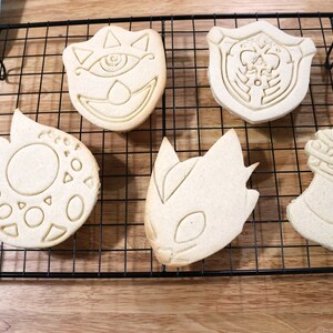 May include: Five plain sugar cookies on a black wire cooling rack. The cookies are shaped like a shield, a sword, a dragon, a creature with an eye, and a circle with geometric shapes.