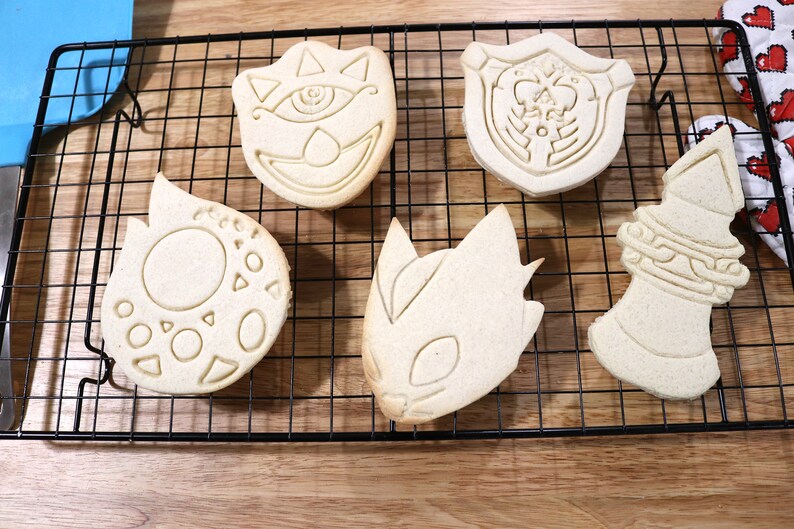 May include: Five plain sugar cookies on a black wire cooling rack. The cookies are shaped like a shield, a sword, a dragon, a creature with an eye, and a circle with geometric shapes.