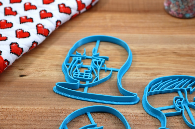 Op de afbeelding: Drie blauwe plastic koekjesvormen in de vorm van gezichten. De vormen liggen op een houten oppervlak.