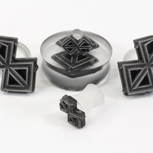 May include: A set of three silver-colored geometric ear gauges. The gauges are in the shape of a cross with a geometric pattern. The gauges are on a white surface.