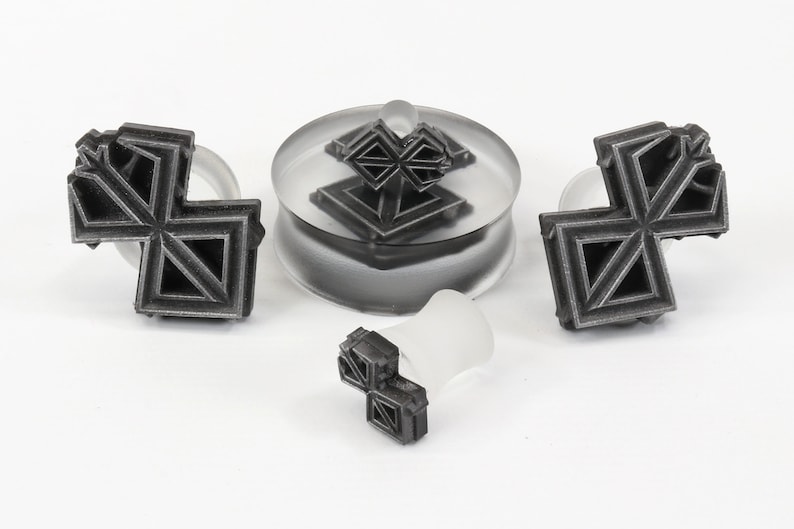 May include: A set of three silver-colored geometric ear gauges. The gauges are in the shape of a cross with a geometric pattern. The gauges are on a white surface.