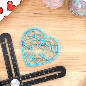May include: A blue plastic heart-shaped cookie cutter with a decorative design. The cutter is on a wooden surface with other baking supplies.