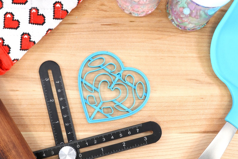 May include: A blue plastic heart-shaped cookie cutter with a decorative design. The cutter is on a wooden surface with other baking supplies.