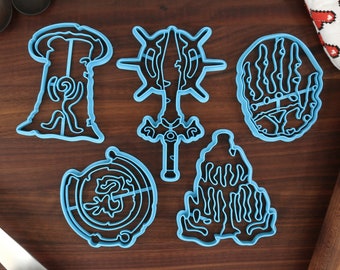Puede incluir: Cinco cortadores de galletas de plástico azul con forma de símbolos de fantasía. Los cortadores están sobre una superficie de madera.