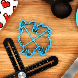 May include: Blue plastic cookie cutter shaped like two dogs standing on their hind legs. The cutter is on a wooden surface with a ruler, a spatula, and a fabric with red hearts.