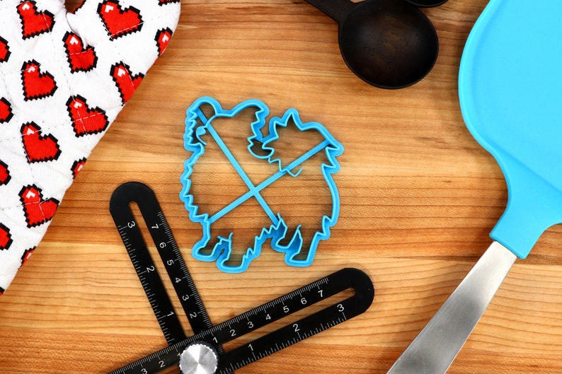May include: Blue plastic cookie cutter shaped like two dogs standing on their hind legs. The cutter is on a wooden surface with a ruler, a spatula, and a fabric with red hearts.