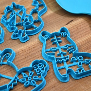 May include: A set of five blue plastic cookie cutters shaped like a tiger, a bunny, a flower, and two other abstract designs. The cutters are on a wooden surface.