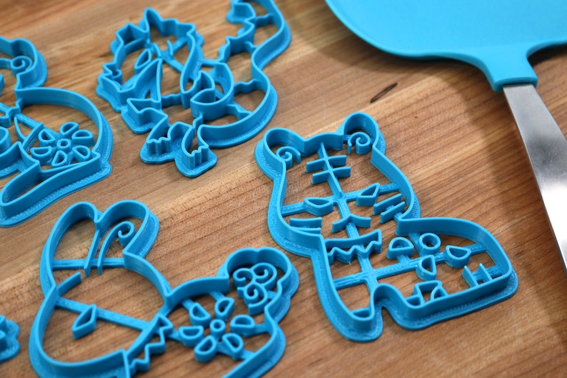 May include: A set of five blue plastic cookie cutters shaped like a tiger, a bunny, a flower, and two other abstract designs. The cutters are on a wooden surface.